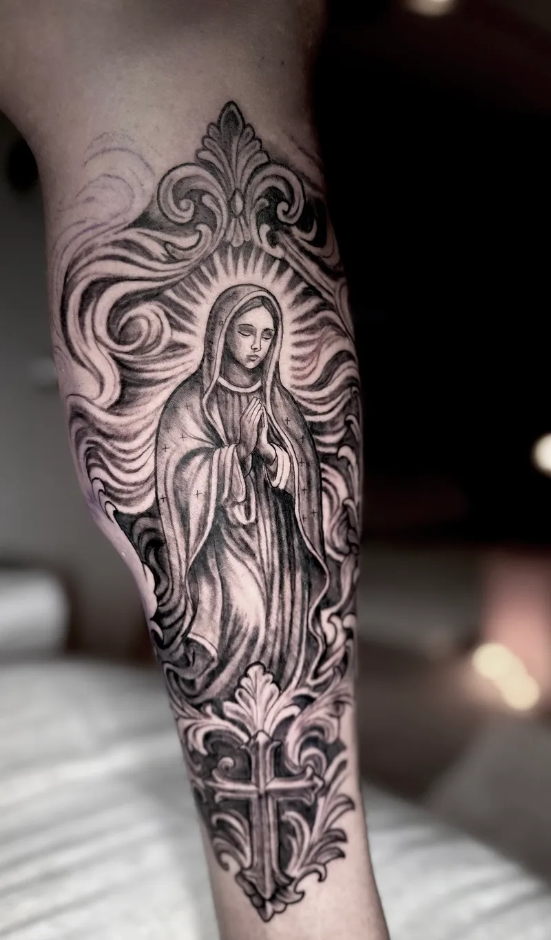 Black and grey tattoo of the Virgin Mary with an ornate frame and cross on the forearm