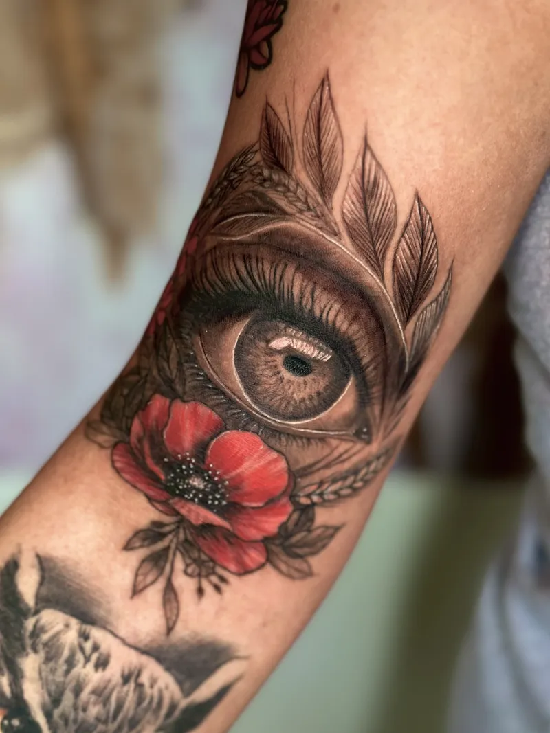 Realism tattoo of an eye with a red poppy and feathers on the forearm