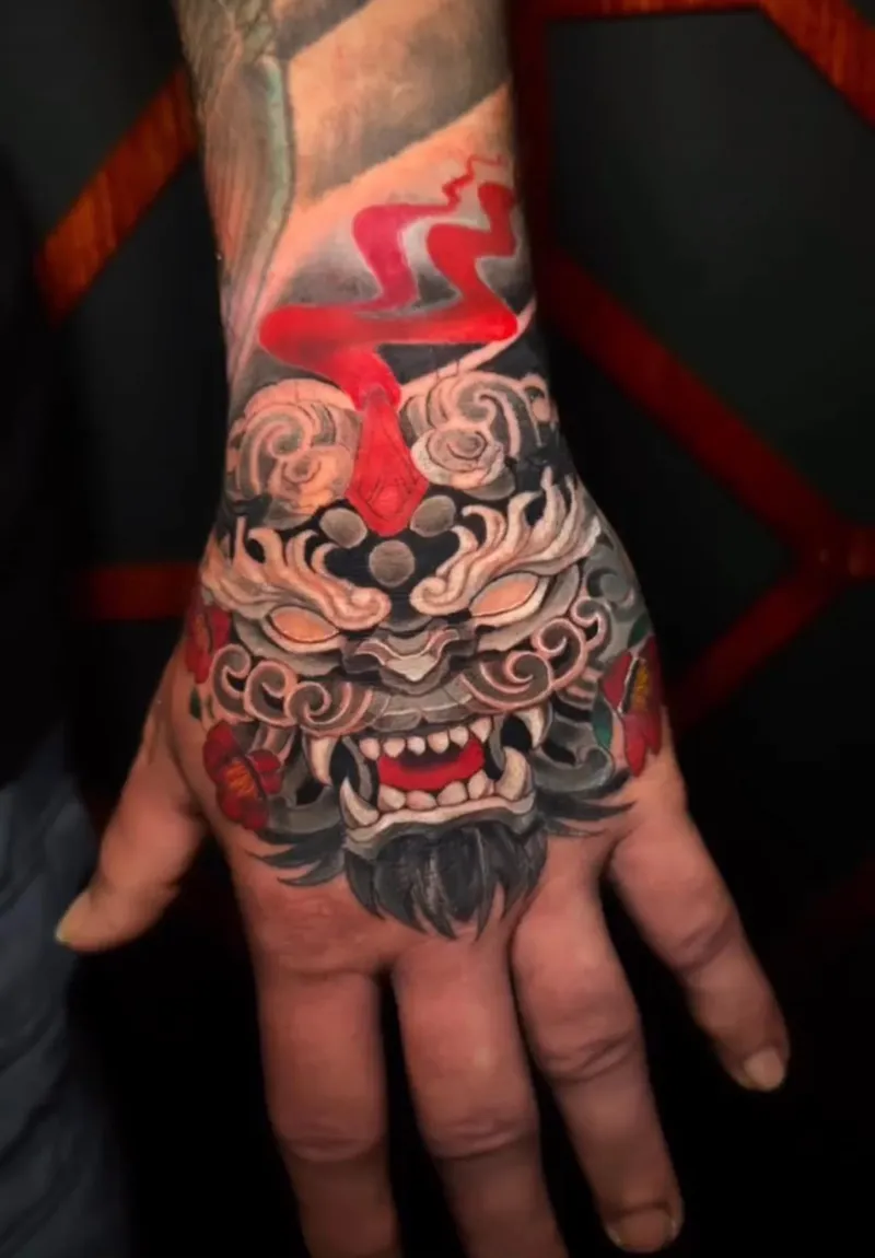 Colour Japanese foo dog tattoo with clouds, red smoke and camellias on the back of the hand, by Nataly Fierro in Omaha, NE